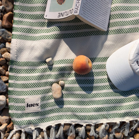 Green and white striped blanket with tassels on pebbles, next to a book and peach.
