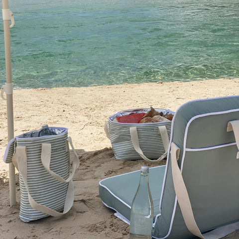 Beach scene with striped bags, cooler, and bottle on sandy shore.