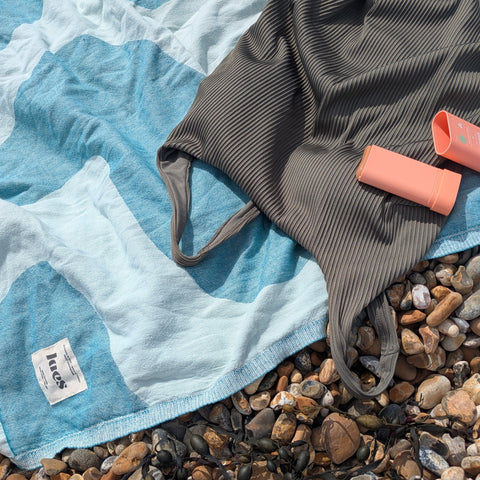 A sage green ribbed swimsuit laid out on a teal and mint patterned beach towel over a pebble beach, with a pink sun protection stick nearby.