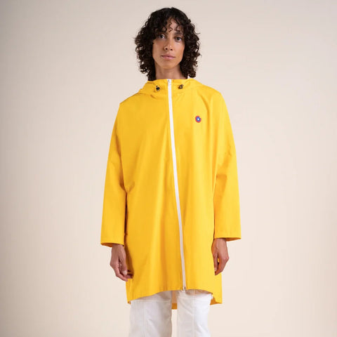 A model wearing a bright yellow hooded raincoat with a white front zipper, paired with white pants and white sneakers, standing against a plain beige studio background.