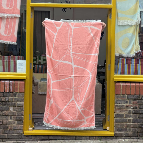 Person holding a pink towel with white patterns in front of a store window displaying various towels.