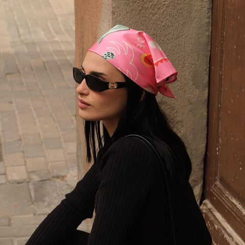 Person wearing a pink headscarf and black sunglasses sitting against a wall.