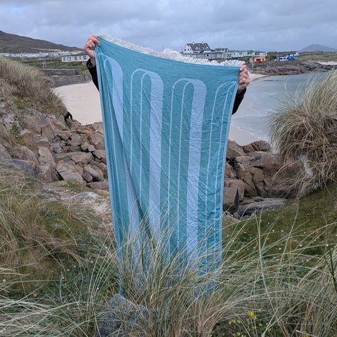 A person outdoors on a rocky beach holding up a large blue towel with vertical light blue stripes.