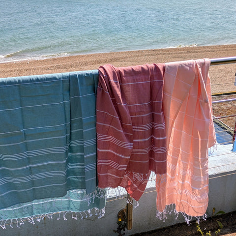 Three beach towels in teal, red and peach hanging on a railing with a sea view.