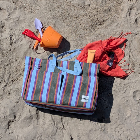 Striped bag with a red towel and orange bucket on sandy beach.