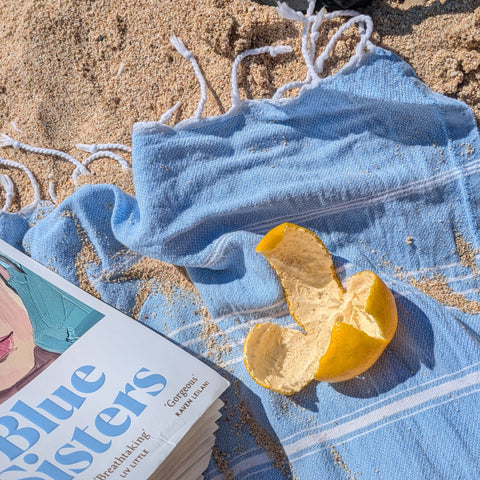 A satsuma peel and a book on a light blue hamman towel on a sandy beach