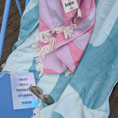 One pink and one teal towel laid over a blue deckchair with sunglasses and a silver book