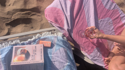 A lifestyle shot of a beach lounge setup featuring a pink and purple patterned beach towel, a blue towel with a book and small orange sunscreen bottle, and a person's hand holding a glass of rose wine on a sandy beach.