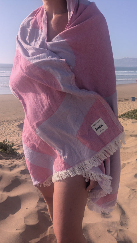 A person wrapped in a pink and lilac abstract patterned beach towel with white fringe, standing on a sandy beach near the ocean.